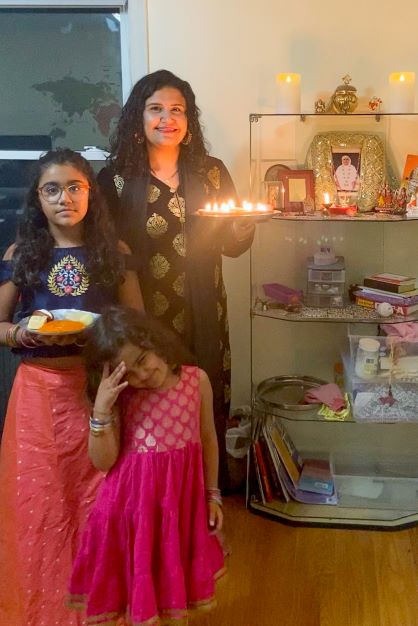 Neha, an Indian woman, and her two young daughters dressed in traditional attire holding candles and dishes