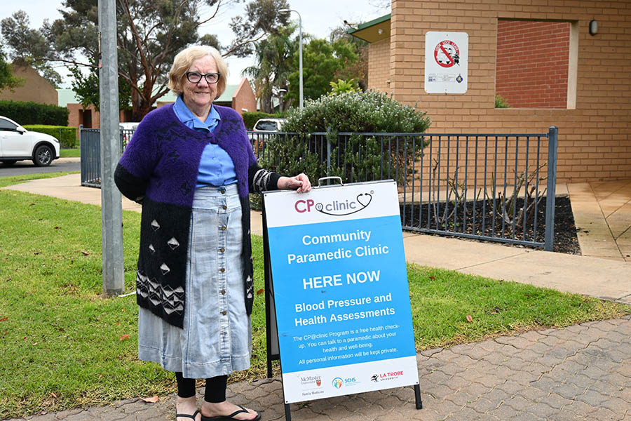 An older adult woman stands beside a sign advertising the CP at clinic program, outside of a community centre.