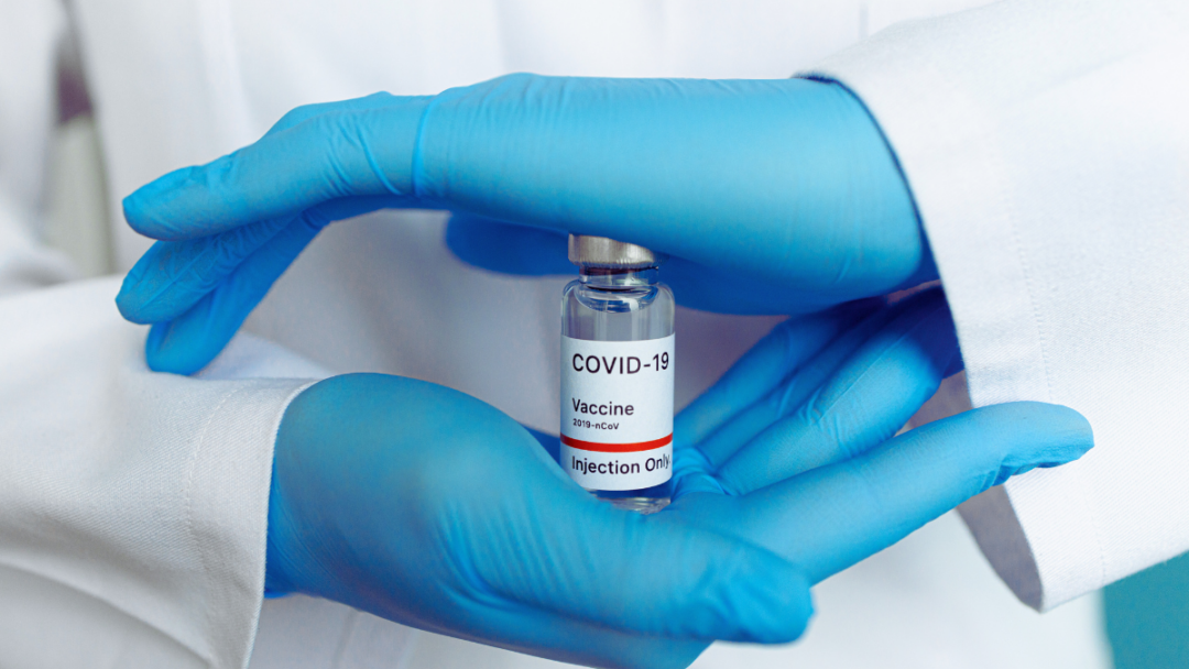 Gloved hands of a person in a lab coat hold a vile of COVID 19 vaccine